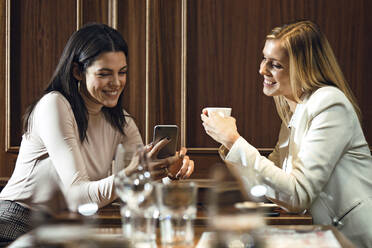 Zwei glückliche Frauen in einem Restaurant, die gemeinsam ein Smartphone betrachten - JSRF00606
