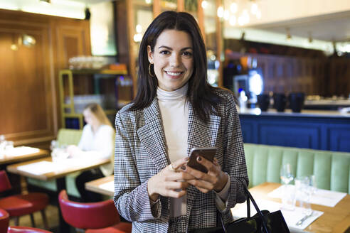 Portrait of smiling businesswoman holding cell phone in a restaurant - JSRF00612