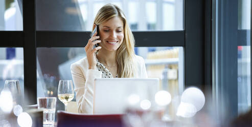 Smiling businesswoman using cell phone and laptop in a restaurant - JSRF00617