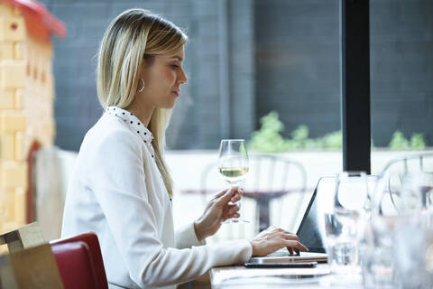 Geschäftsfrau mit Laptop in einem Restaurant, während sie Weißwein trinkt, lizenzfreies Stockfoto