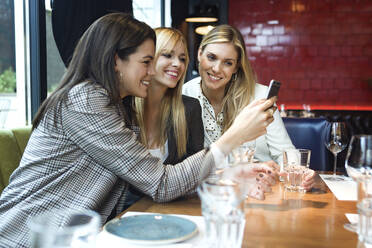 Drei glückliche Geschäftsfrauen in einem Restaurant, die gemeinsam ein Smartphone betrachten - JSRF00628