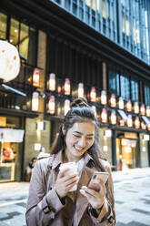 Glückliche junge Frau hält ein Getränk zum Mitnehmen in der Hand und benutzt ein Smartphone in Ginza, Tokio, Japan - MCVF00027