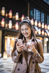 Glückliche junge Frau hält ein Getränk zum Mitnehmen in der Hand und benutzt ein Smartphone in Ginza, Tokio, Japan - MCVF00030