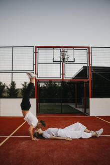 Junges Paar auf einem Basketballplatz, Frau liegt und Mann macht einen Handstand und küsst sie - LHPF00883