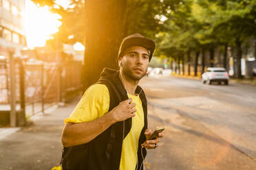 Porträt eines jungen Mannes, der mit Kopfhörern und Smartphone Musik hört und am Straßenrand wartet - GIOF07148