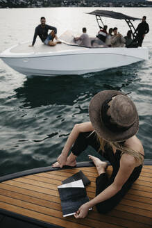 Frau mit einem Buch auf einem Boot mit Blick auf ein Boot mit jungen Männern - LHPF00934