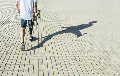 Junger Mann mit Beinprothese geht und hält Skateboard, lizenzfreies Stockfoto