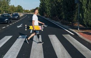 Junger Mann mit Beinprothese hält Skateboard und geht auf dem Zebrastreifen - JCMF00227