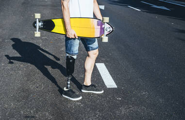 Junger Mann mit Beinprothese hält Skateboard auf einer Straße - JCMF00228