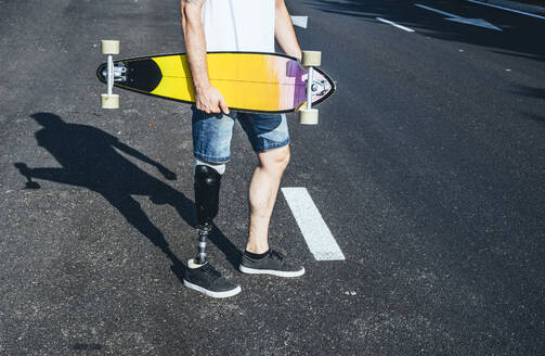 Junger Mann mit Beinprothese hält Skateboard auf einer Straße - JCMF00228