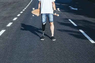 Junger Mann mit Beinprothese hält Skateboard auf einer Straße - JCMF00229