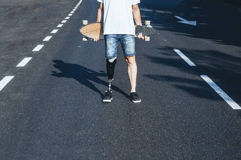 Junger Mann mit Beinprothese hält Skateboard auf einer Straße - JCMF00229