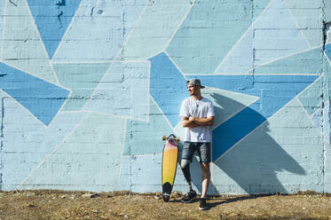 Young man with leg prosthesis standing against concrete wall with a skateboard - JCMF00230