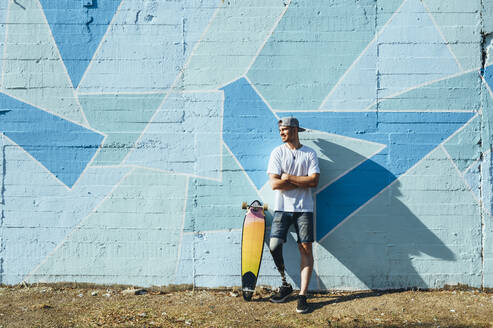 Junger Mann mit Beinprothese steht mit einem Skateboard an einer Betonwand - JCMF00230