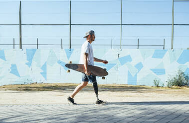 Junger Mann mit Beinprothese geht und hält Skateboard - JCMF00231