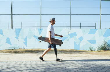 Young man with leg prosthesis walking and holding skateboard - JCMF00231