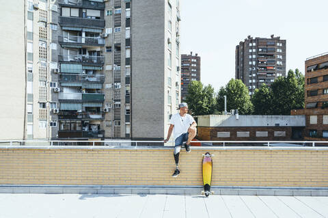 Junger Mann mit Beinprothese sitzt auf einem Geländer in der Stadt neben einem Skateboard, lizenzfreies Stockfoto