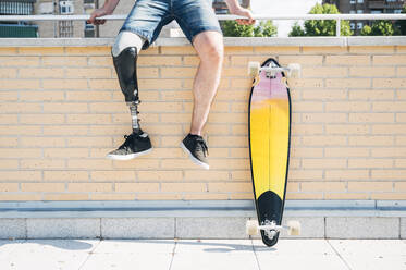 Junger Mann mit Beinprothese sitzt auf einem Geländer in der Stadt neben einem Skateboard - JCMF00235