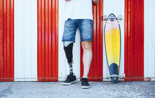 Young man with leg prosthesis standing next to skateboard at a wall - JCMF00236