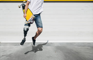 Young man with leg prosthesis holding skateboard and jumping - JCMF00238