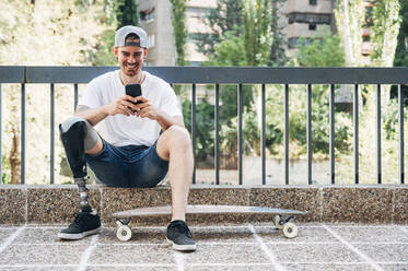 Lächelnder junger Mann mit Beinprothese und Skateboard, der ein Smartphone benutzt - JCMF00239