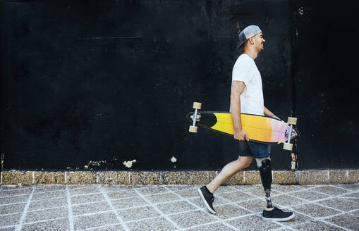 Young man with leg prosthesis holding skateboard passing a wall - JCMF00241