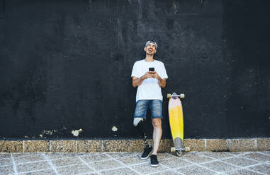 Lachender junger Mann mit Beinprothese und Skateboard, der ein Smartphone benutzt - JCMF00242