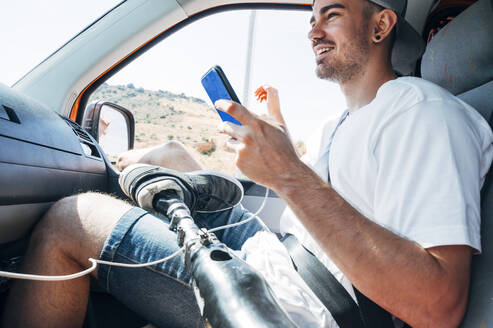 Lächelnder junger Mann mit Beinprothese, der im Wohnmobil sitzt und sein Smartphone benutzt - JCMF00243