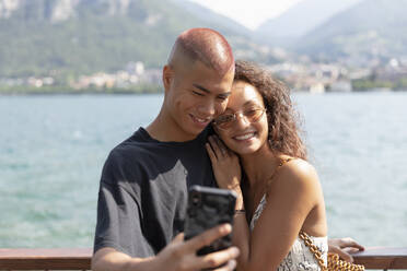 Porträt eines glücklichen jungen Paares, das ein Selfie vor dem Comer See macht, Lecco, Italien - MCVF00047