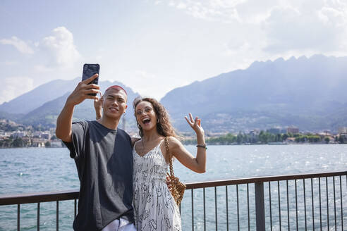 Porträt eines jungen Paares, das ein Selfie vor dem Comer See macht, Lecco, Italien - MCVF00050