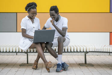 Smiling young couple sitting on bench looking at laptop - DLTSF00185