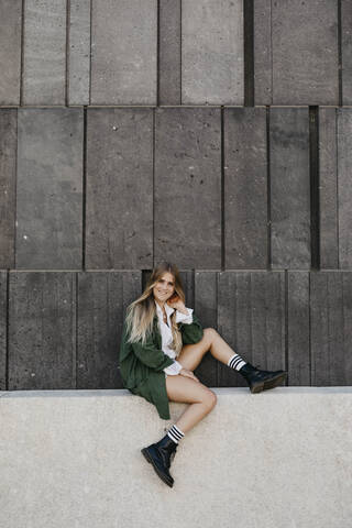 Portrait of blond young woman wearing green jacket sitting on a wall, Vienna, Austria stock photo