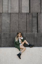 Portrait of blond young woman wearing green jacket sitting on a wall, Vienna, Austria - LHPF00956