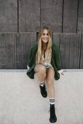 Portrait of smiling blond woman wearing green jacket sitting on a wall, Vienna, Austria - LHPF00957