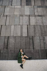 Blonde junge Frau mit grüner Jacke sitzt auf einer Mauer, Wien, Österreich - LHPF00958