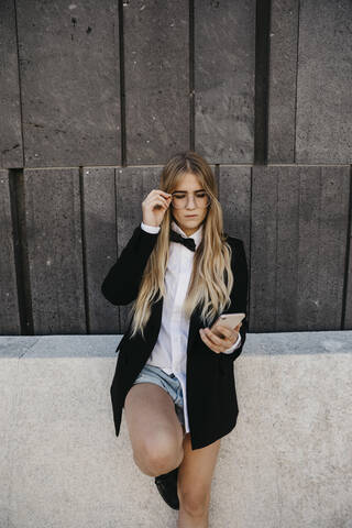 Porträt einer blonden jungen Frau mit schwarzer Krawatte und Blazer, die auf ihr Mobiltelefon schaut, Wien, Österreich, lizenzfreies Stockfoto