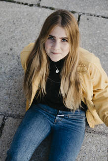 Porträt einer lächelnden jungen Frau mit gelber Jacke, die auf dem Boden sitzt - LHPF00971