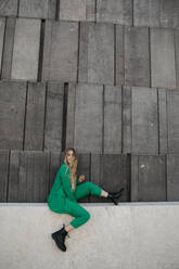 Blond young woman wearing green pantsuit sitting on a wall, Vienna, Austria - LHPF00980