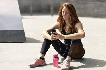 Sporty young woman with earphones having a break using smartphone outdoors - JSMF01292