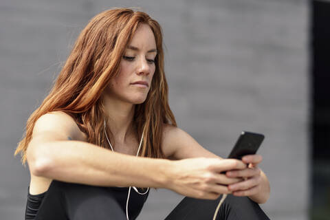 Sportliche junge Frau mit Kopfhörern macht eine Pause und benutzt ihr Smartphone im Freien, lizenzfreies Stockfoto