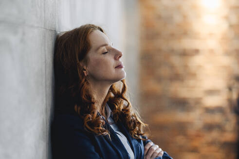 Redheaded businesswoman having a break leaning against a wall in office - KNSF06594