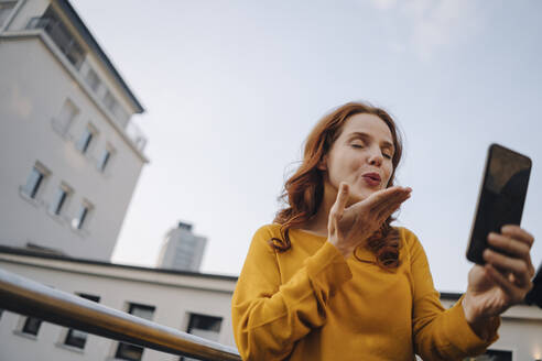 Redheaded woman using cell phone on roof terrace blowing a kiss - KNSF06621