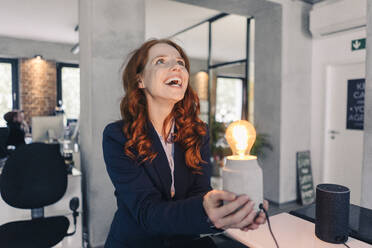 Laughing redheaded businesswoman holding lamp in office - KNSF06641