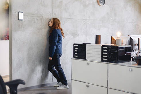Tired businesswoman leaning against a wall in office - KNSF06642