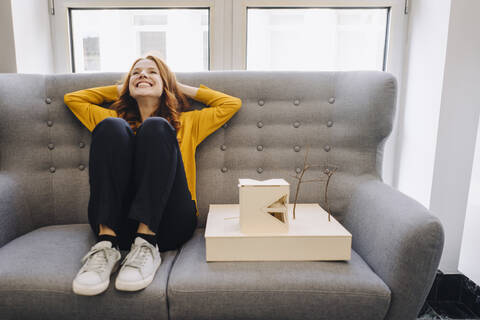 Glückliche Frau sitzt auf einer Couch neben einem Architekturmodell, lizenzfreies Stockfoto