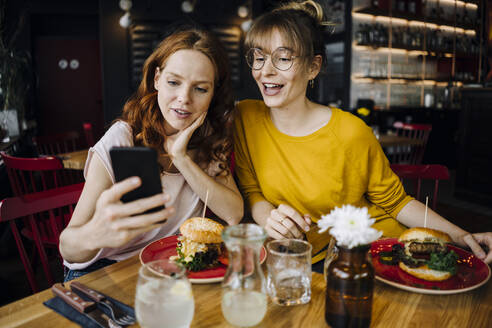 Two female friends with cell phone having burger in a restaurant - KNSF06659