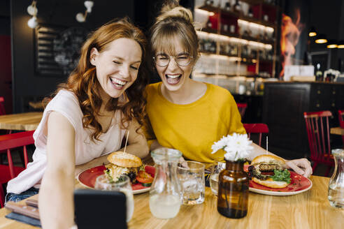 Zwei glückliche Freundinnen essen einen Burger und machen ein Selfie in einem Restaurant - KNSF06660