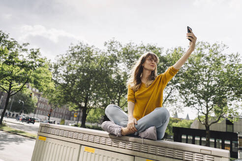 Junge Frau sitzt auf einer Kiste in der Stadt und macht ein Selfie - KNSF06673