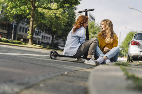 Female friends with e-scooter talking at the roadside in the city - KNSF06687