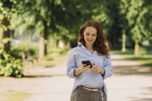 Lächelnde rothaarige Frau benutzt Handy im Park - KNSF06727